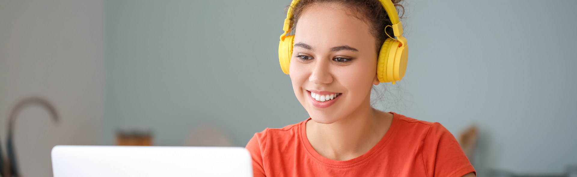 A woman smiling, wearing yellow headphones and looking at a computer screen.