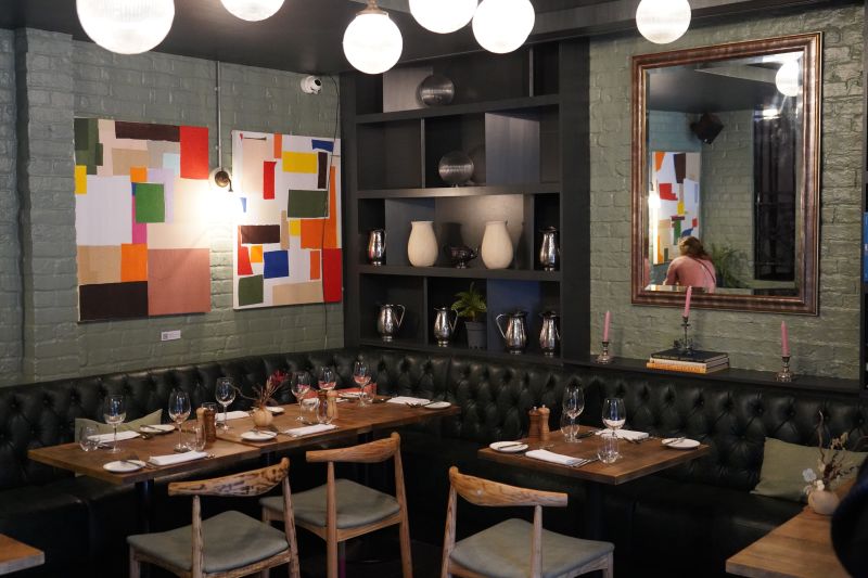 The image shows a cosy fine-dining restaurant interior with green-painted brick walls and modern decor. A tufted green leather bench lines the wall, paired with wooden tables set for guests, including wine glasses, plates, and utensils. The space features vibrant abstract artwork on the wall, alongside shelves. A large mirror adds depth to the room, reflecting part of the restaurant's ambiance, while warm lighting from globe-shaped fixtures enhances the space.