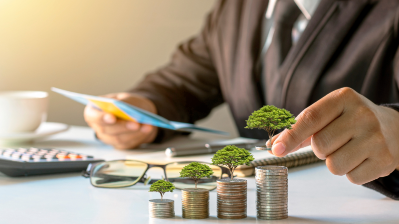 A business professional in a suit is seated at a desk, holding documents. In the foreground, stacks of coins gradually increase in height, with each stack featuring a small tree growing on top. A calculator, glasses, and a cup of coffee are also visible on the desk.